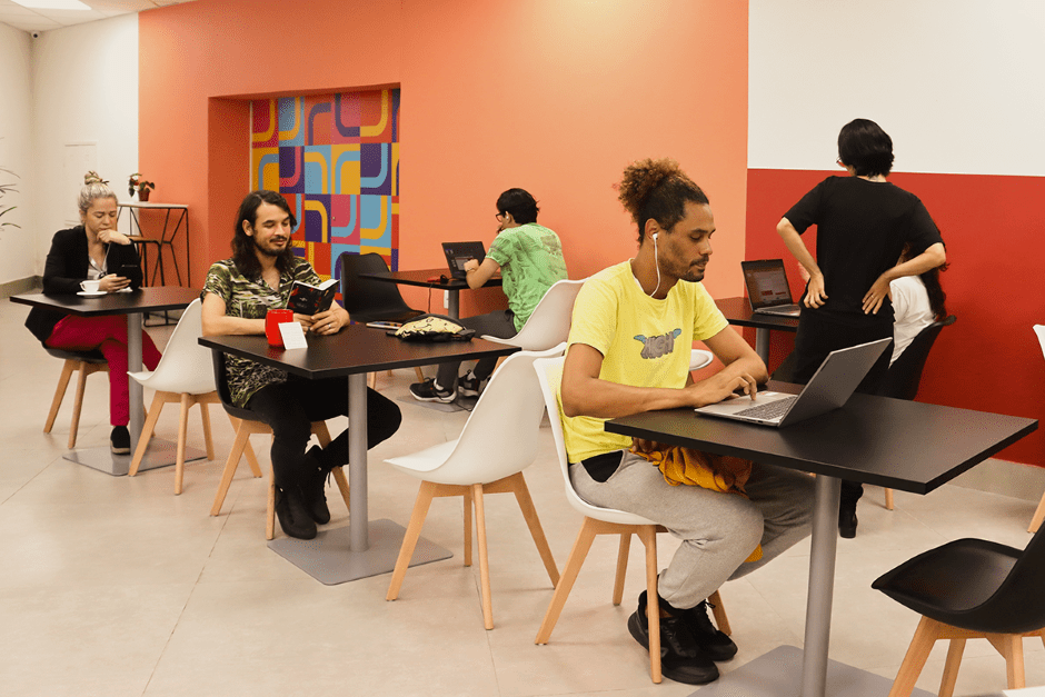 Grupo diverso de pessoas sentado em mesas individuais no espaço do Lab Café no Hub ES +, utilizando notebooks para trabalho ou estudo.